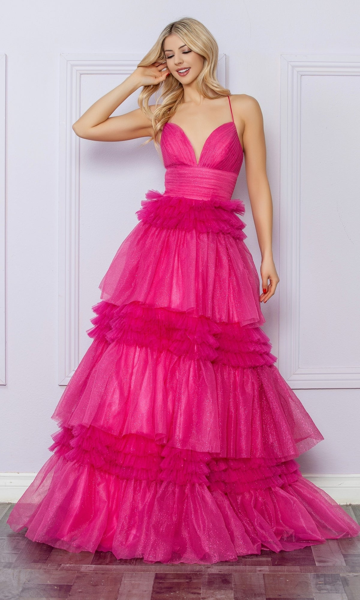 A woman stands indoors, smiling as she touches her hair, wearing the Nox Anabel Ruffled Glitter Prom Dress R1316—a bright pink gown with a tiered tulle skirt, deep V-neckline, and spaghetti straps.
